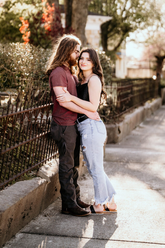 Male standing with his back to a metal fence while his fiancé leans into him embracing each other