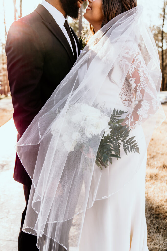 Bride and groom nuzzle close while the glow of the sun swarms around them. The brides veil is draped over her shoulder and her bouquet