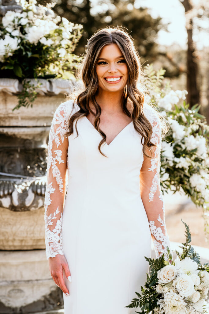 The bride standing smiling in front of the floral water fountain lit by the glow of the sun with her bouquet dropped by her hip