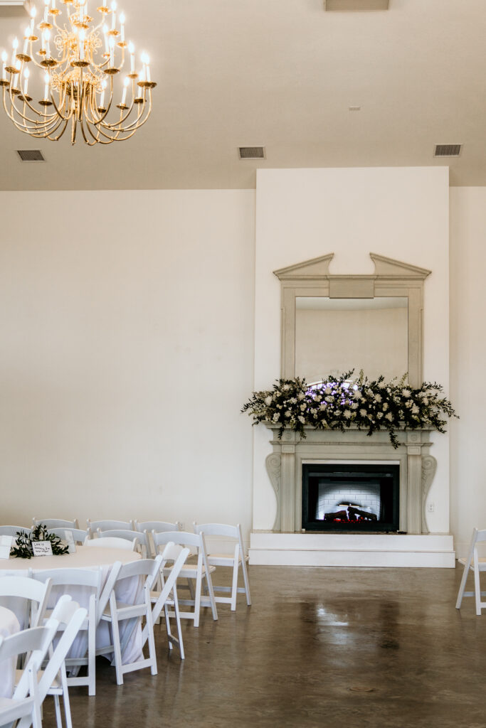 Grand fireplace in the center of the room decorated with florals and a large chandelier hanging from the ceiling of the grand room set up with white tables and chairs for a wedding.