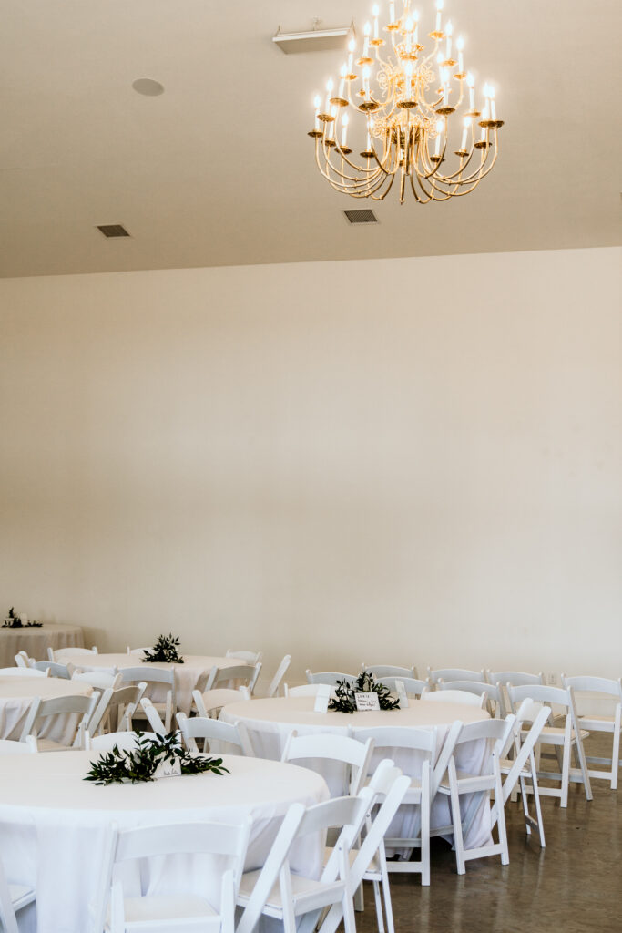 Large chandelier hanging from the ceiling over tables with white linens and white chairs for the reception