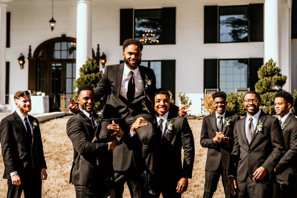 Groom being carried by groomsmen toward the camera in front of the wedding venue