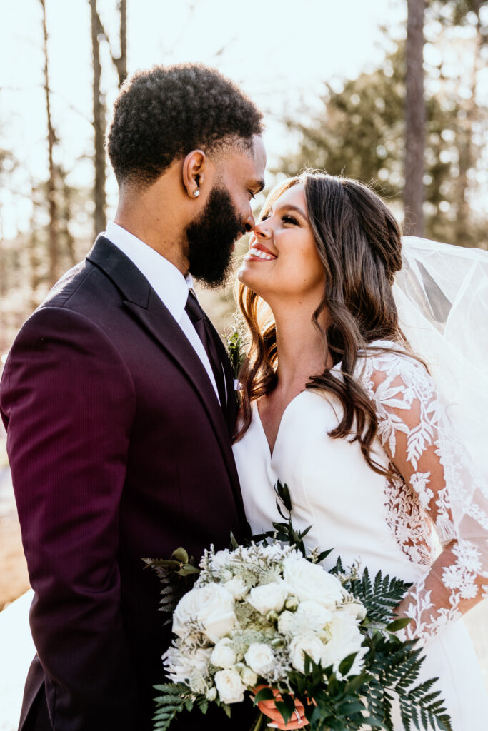 Bride and groom nose-to-nose while smiling at each other. The bride holds her bouquet at her side while her veil flies behind her