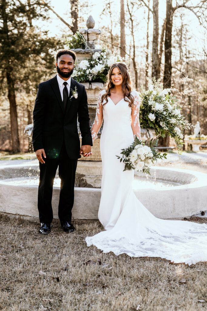 Bride and groom are holding hands in front of the water fountain decorated with white blooms and greenery while the bride holds her bouquet at her side lit by the glow of sunset