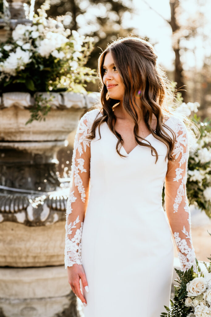 The bride standing in front of the water fountain back lit by the sunset with both arms at her side while she looks off to the side at something in the distance