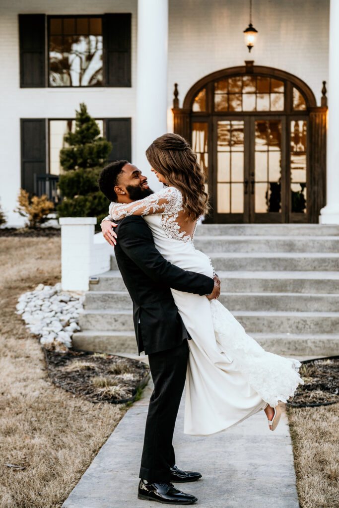 Groom picks up and spins his bride on the side walk leading to the front entrance of their wedding venue